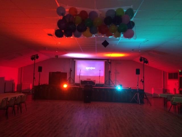 Salle décorée de ballons avec écran et installation DJ éclairée en rouge et cyan.