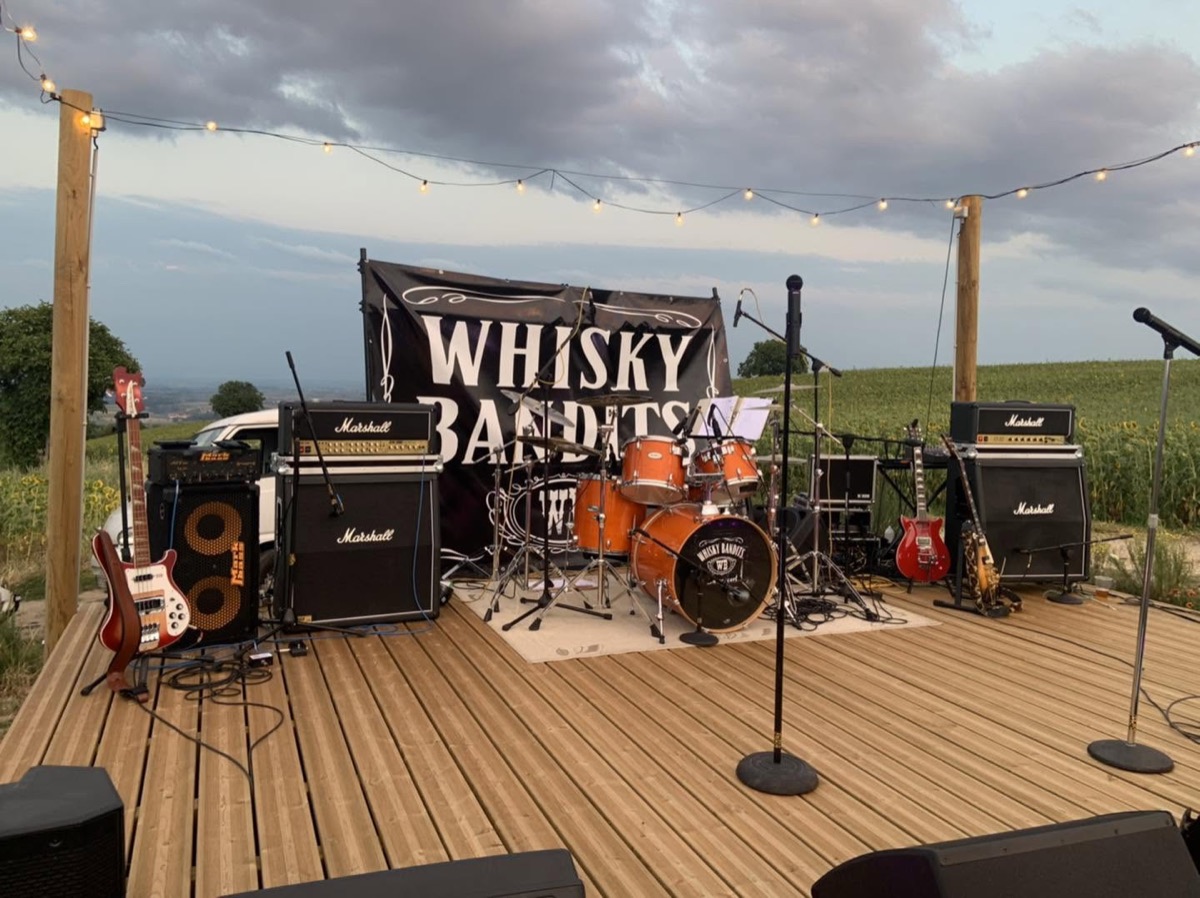Scène rock en extérieur avec batterie, guitares, amplis, micros et retours de scène.